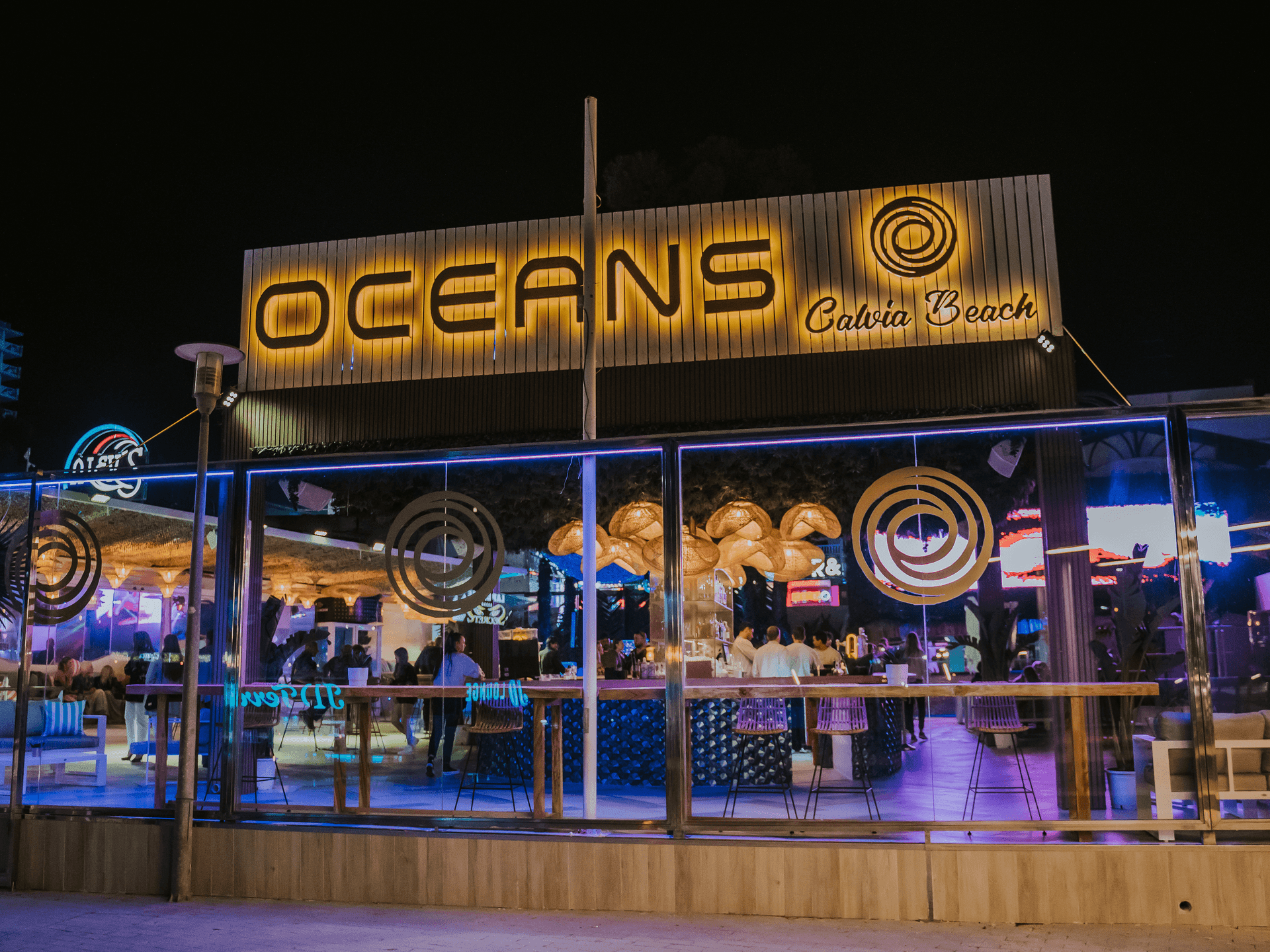 Exterior view of Oceans Calvia Beach restaurant illuminated at night, with people dining inside.